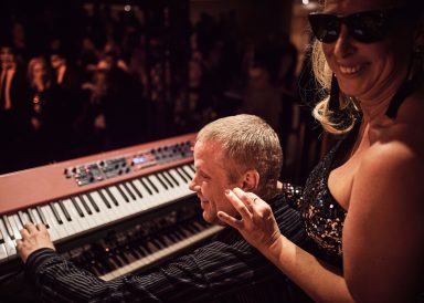 Ein Musiker spielt auf einem Keyboard, während eine Frau ihm freundlich zusieht.