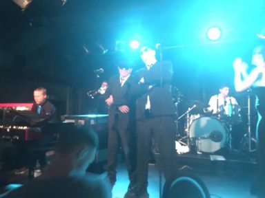 Zwei Musiker in Anzügen auf einer Bühne mit Band, blaues Licht im Hintergrund.