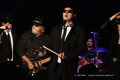Sechs Musiker in schwarzen Anzügen und Sonnenbrillen, live auf der Bühne.