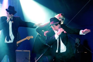 Drei Musiker in Anzügen und Sonnenbrillen performen auf einer Bühnenshow.