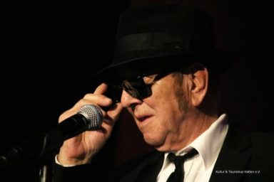 Mann in schwarzem Hut und Sonnenbrille, singt ins Mikrofon.