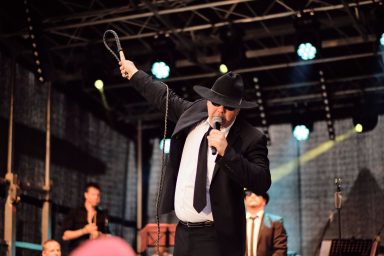 Mann in Anzug und Hut präsentiert eine Show auf einer Bühne mit Lichtern.