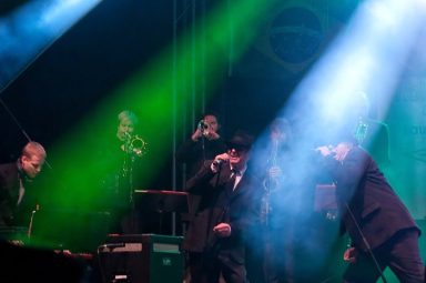 Musikgruppe auf der Bühne, umgeben von bunten Lichtstrahlen in verschiedenen Farben.