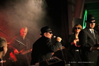 Musiker auf der Bühne, in Schwarz gekleidet, mit Rauch und rotem Licht im Hintergrund.