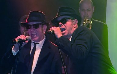 Zwei Männer in Anzügen und Sonnenbrillen singen auf einer Bühne.