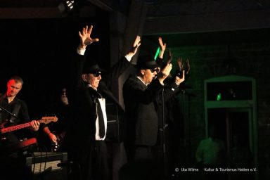Musiker in Anzügen geben eine Live-Performance mit gestreckten Armen auf der Bühne.