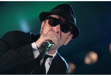 Mann in Anzug und Hut singt ins Mikrofon, trägt Sonnenbrille, Bühnenbeleuchtung im Hintergrund.