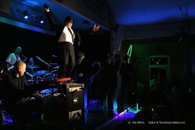 Ein Musiker führt auf einer Bühne mit Band auf, umgeben von buntem Licht.