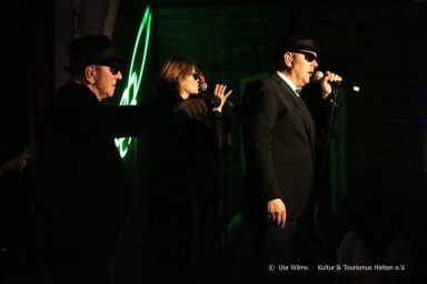 Drei Musiker in schwarzer Kleidung, mit Sonnenbrillen, performen auf der Bühne.