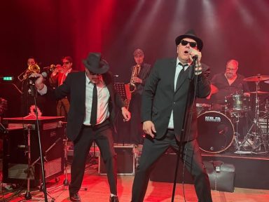 Zwei Sänger in schwarzer Kleidung performen auf einer Bühne mit Band im Hintergrund.