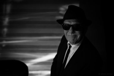 Mann in Anzug und Hut, trägt Sonnenbrille, lächelt in einer kontrastreichen Umgebung.