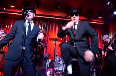 Zwei Musiker in Anzügen und Sonnenbrillen treten auf einer Bühne mit Band auf.