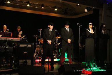 Eine live Band mit fünf Mitgliedern auf der Bühne, in schwarzer Kleidung und Sonnenbrillen.