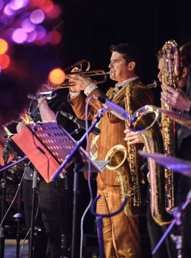 Ein Musiker spielt Trompete, umgeben von Saxophonisten auf einer Bühne.