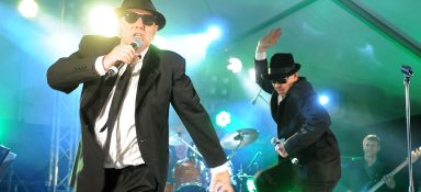 Zwei Musiker in Anzügen performen auf einer Bühne mit bunten Lichtern im Hintergrund.
