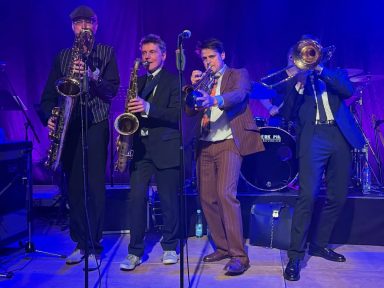 Vier Musiker mit Blasinstrumenten spielen auf einer Bühne vor lila Hintergrund.
