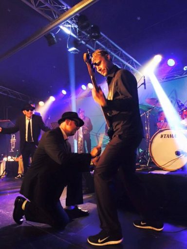 Zwei Musiker auf einer Bühnenaufführung, einer spielt Gitarre, der andere kniet daneben.