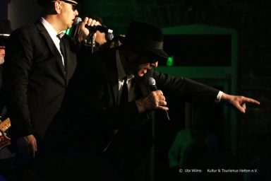 Zwei Musiker in Anzügen und Hüten singen auf einer Bühne mit grünem Licht.