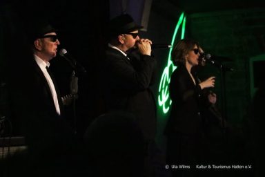 Drei Musiker in Anzügen und Sonnenbrillen singen auf einer Bühne.
