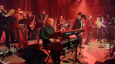 Eine Band mit Musikern, die auf einer Bühne live performen. Rote Beleuchtung im Hintergrund.