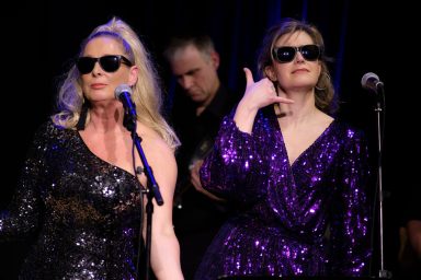 Zwei Sängerinnen in Sonnenbrillen, glitzernden Outfits, performen auf der Bühne.