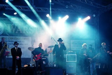 Band auf der Bühne mit Instrumenten, umgeben von bunten Lichtern und Nebel.
