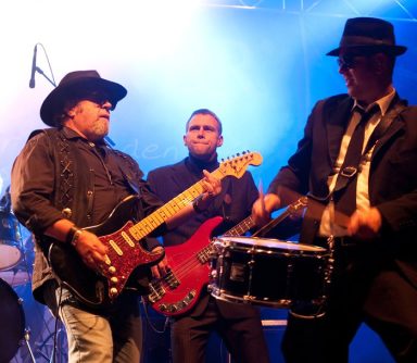 Drei Musiker auf der Bühne, einer mit Gitarre, einer mit Trommel, dunkle Anzüge und Hüte.