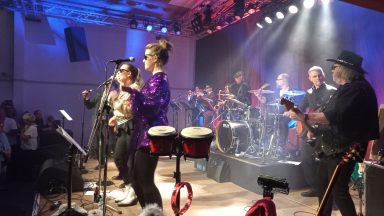 Band spielt auf einer Bühne, mit Musikern, die verschiedene Instrumente spielen.