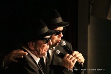 Zwei Männer in Anzügen und Hüten singen ins Mikrofon.
