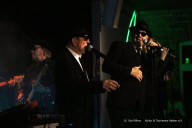 Zwei Sänger in schwarzer Kleidung, die auf einer Bühne performen. Musiker im Hintergrund.