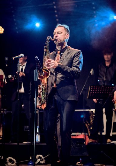 Mann mit Saxophon auf der Bühne, umgeben von Musikern in festlicher Stimmung.