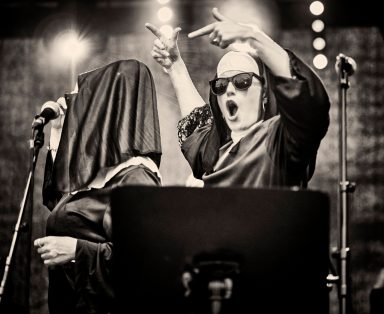 Zwei Performerinnen auf der Bühne, eine mit Kopftuch und die andere in Sonnenbrille, feiern.