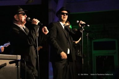 Zwei Musiker in Anzügen und Sonnenbrillen singen in ein Mikrofon auf einer Bühne.