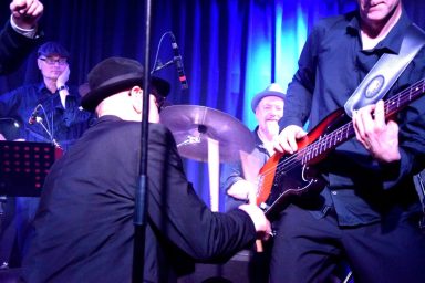 Eine Band spielt live auf der Bühne, Musiker in der Nähe von Instrumenten, lebendige Lichtstimmung.