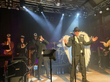 Eine Band mit einem Sänger in Anzug und Flügeln, umgeben von Musiker:innen in Kostümen.
