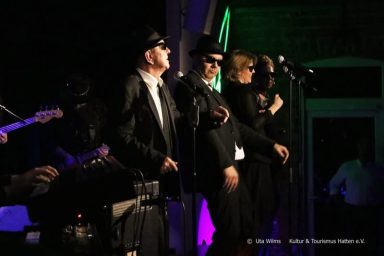 Drei Sänger in Schwarz mit Sonnenbrillen stehen auf einer Bühne und performen.