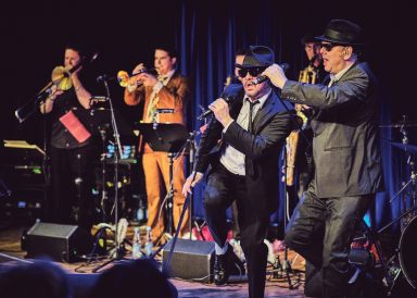 Band mit drei Sängern in Anzügen, die auf der Bühne performen, umgeben von Musikern.