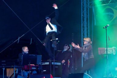 Ein Musiker springt auf der Bühne, während andere Bandmitglieder ihn anfeuern.