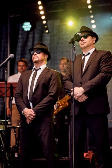 Zwei Männer in Anzügen und Sonnenbrillen stehen auf der Bühne und singen.
