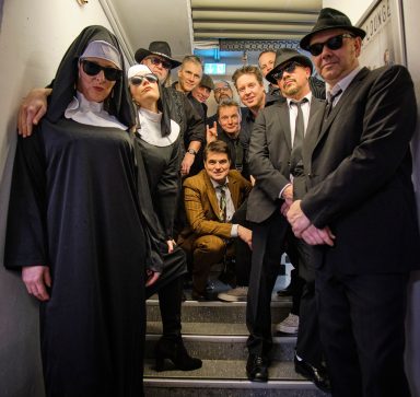 Gruppe von Personen in Anzügen und Sonnenbrillen, zwei im Nonnenkostüm, auf einer Treppe.