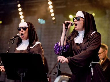 Zwei Nonnen mit Sonnenbrillen singen in Mikrofone, umgeben von einer Band.