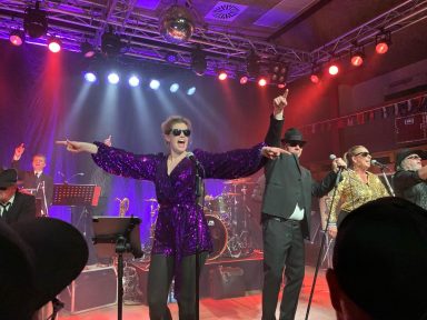 Ein Musiker in lila Outfit und Sonnenbrille, umgeben von Bandkollegen auf der Bühne.