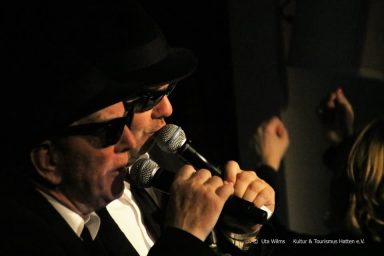 Zwei Sänger in Anzügen und Sonnenbrillen halten Mikrofone und singen gemeinsam.