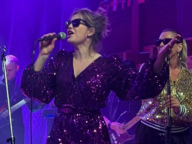 Sängerin mit Sonnenbrille in glitzerndem Outfit, umgeben von Bandmitgliedern auf der Bühne.