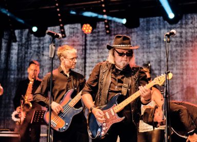 Musiker spielen auf der Bühne, einer mit Sonnenbrille und E-Gitarre, Gruppenatmosphäre.