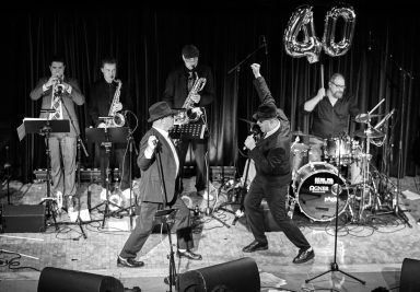 Zwei Sänger performen leidenschaftlich vor einer Band mit Trompete und Schlagzeug.
