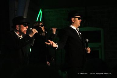Drei Männer in Anzügen und Sonnenbrillen performen auf einer Bühne.
