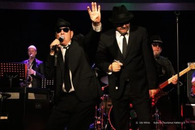 Zwei Sänger in Anzügen und Sonnenbrillen performen auf einer Bühnenveranstaltung.