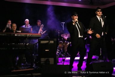 Zwei Sänger in Anzügen performen auf einer Bühne, begleitet von einer Band und bunten Lichtern.