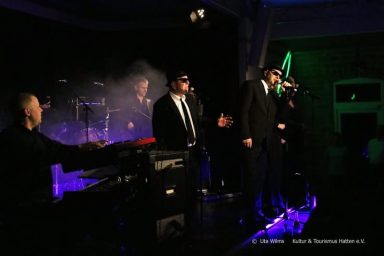 Eine Band in Anzügen performt auf der Bühne mit Lichtern und Nebel.